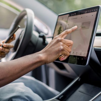 Autopilot self-driving car system with no human intervention. Close up cropped image of hand of African male driver browsing the internet using smartphone and touchscreen in futuristic autonomous car.